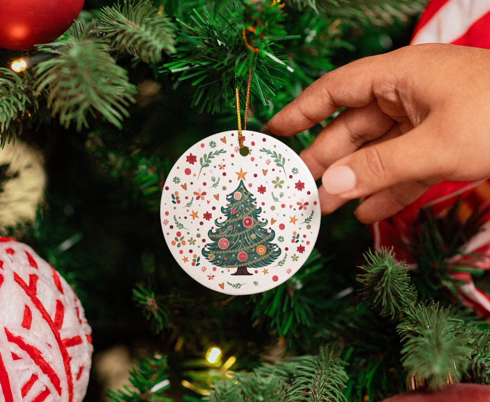 Hand holding a decorative Christmas ornament with a tree design against a Christmas tree background.